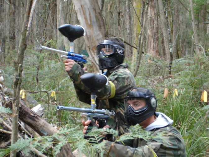 Two paintball players hiding behind a tree aiming at their targets.